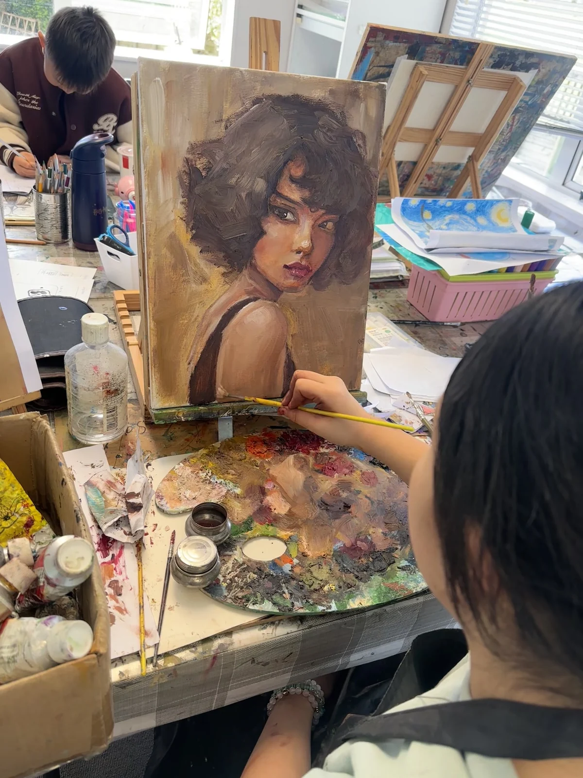 An older learner painting a warm-toned portrait at a busy studio table