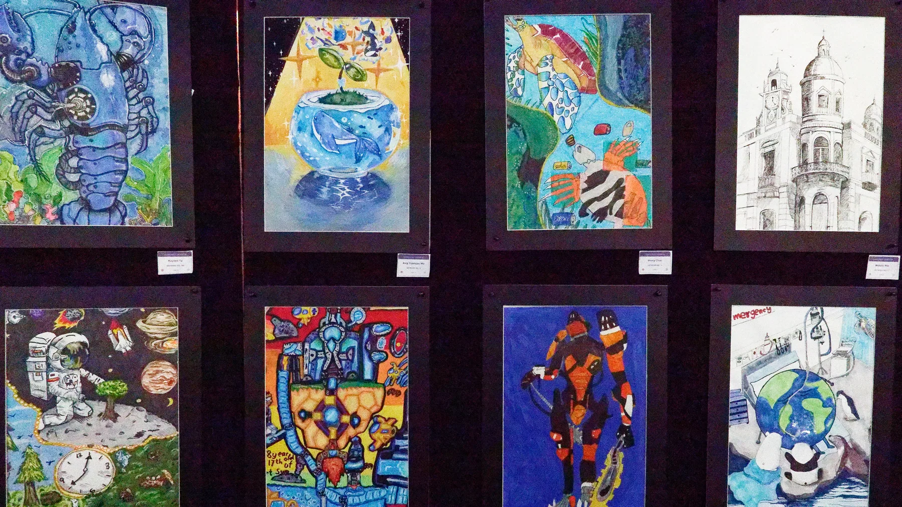 Framed student paintings and drawings displayed together on a dark exhibition wall